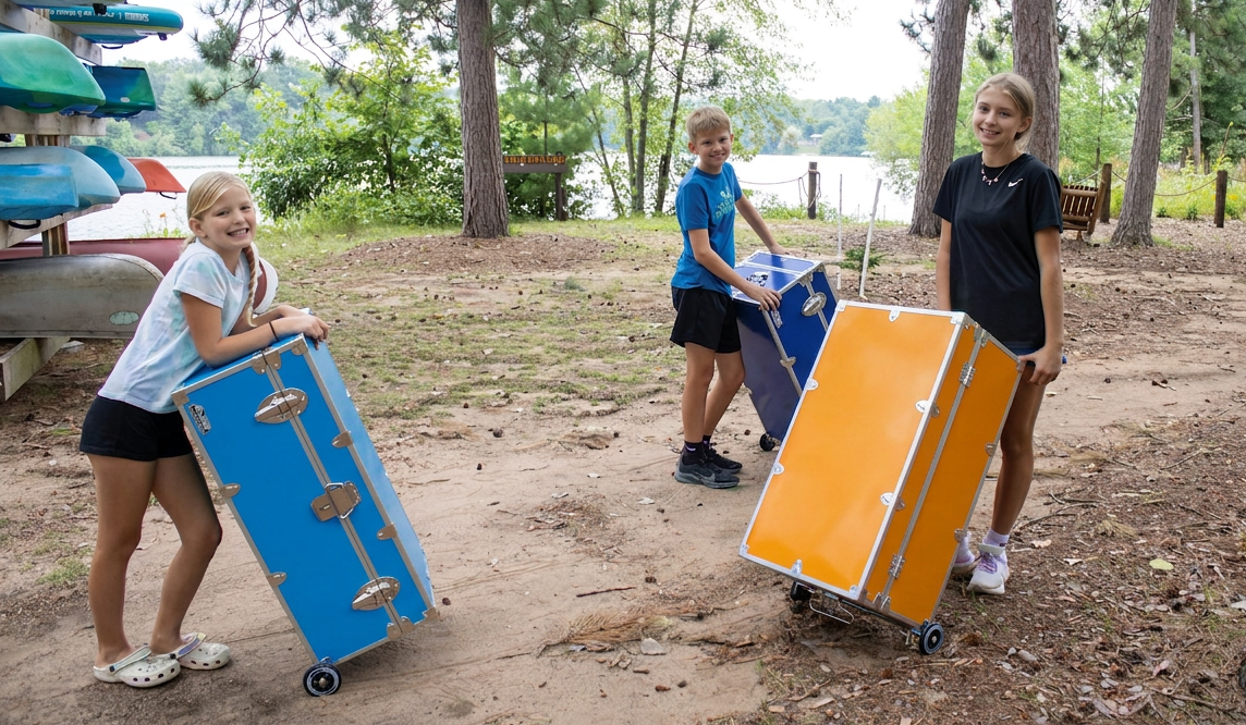 Three children pushing colorful folding chairs outdoors near a lake.
