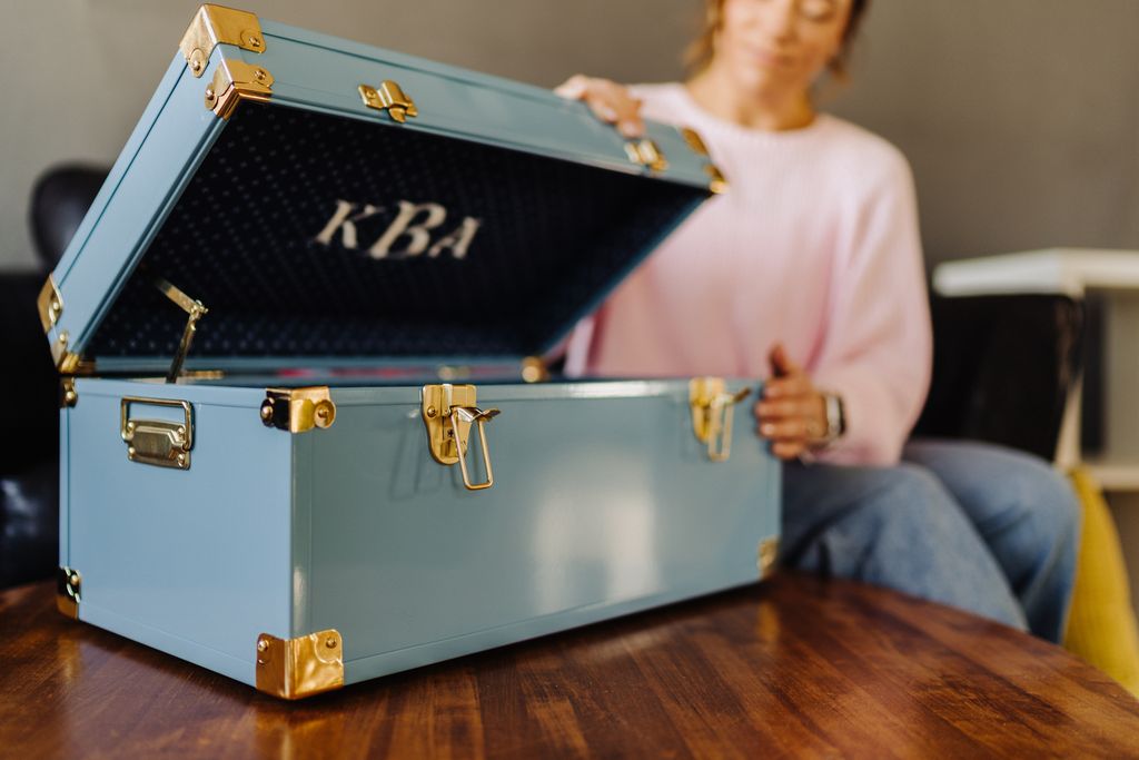 Person opening a blue trunk with gold accents on a wooden table, featuring the initials 'KBA'.