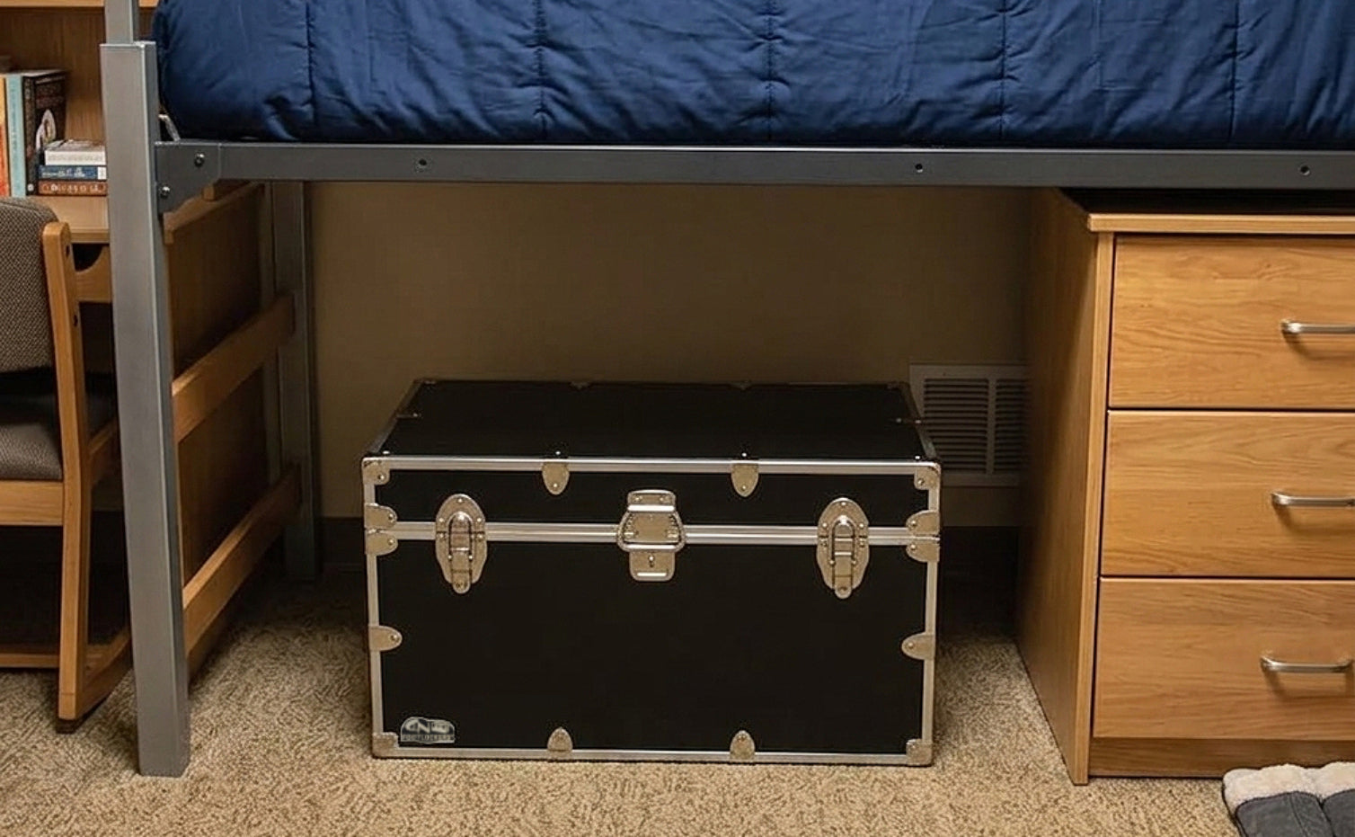 Black trunk with silver latches on a carpeted floor next to a bed and wooden furniture.