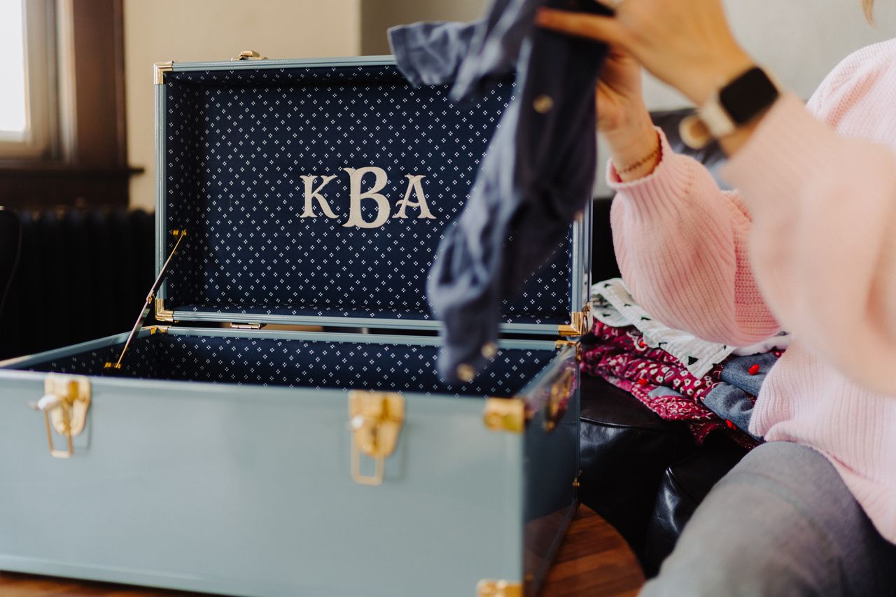 Person sorting keepsakes with clothes, monogrammed 'KBA' on the trunk lid.